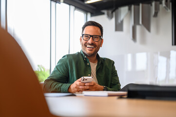 Confident project manager in glasses using smartphone and looking away while sitting at desk in office