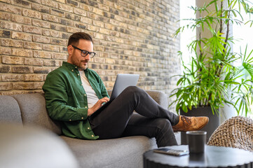 Male computer programmer with glasses working over wireless computer while sitting on sofa in office
