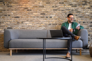 Handsome employee holding coffee cup and using social media apps over smart phone on sofa in office