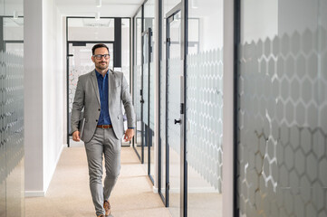 Full length of handsome young financial advisor holding computer and walking in office corridor