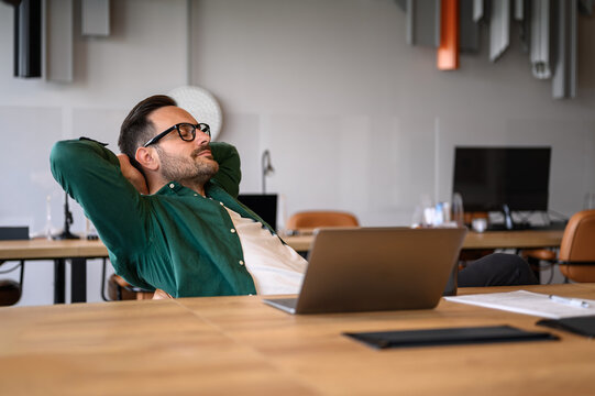 Tired young businessman with hands behind head and eyes closed taking a break at his desk in office