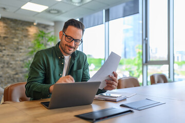 Handsome IT engineer in eyeglasses smiling and developing online website over laptop on desk in...