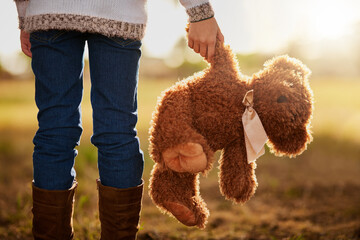 Teddy bear, legs and back of child in nature for support, fantasy and memory in field. Animal or...