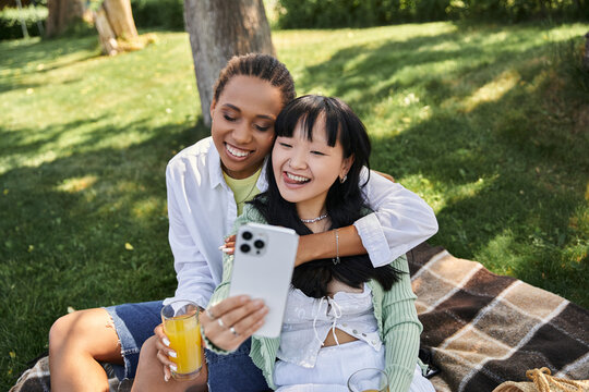 Two women, an African American and an Asian, enjoy a picnic in the park, laughing and taking a selfie. - Powered by Adobe