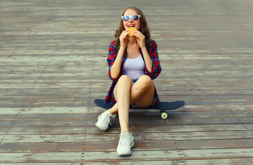 Happy modern young woman eating burger fast food, stylish teenage girl sitting on skateboard in city