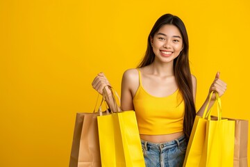 Cheerful young Asian woman in yellow sleeveless top holding shopping bags with a thumbs-up on a yellow background, concept of consumerism and retail joy