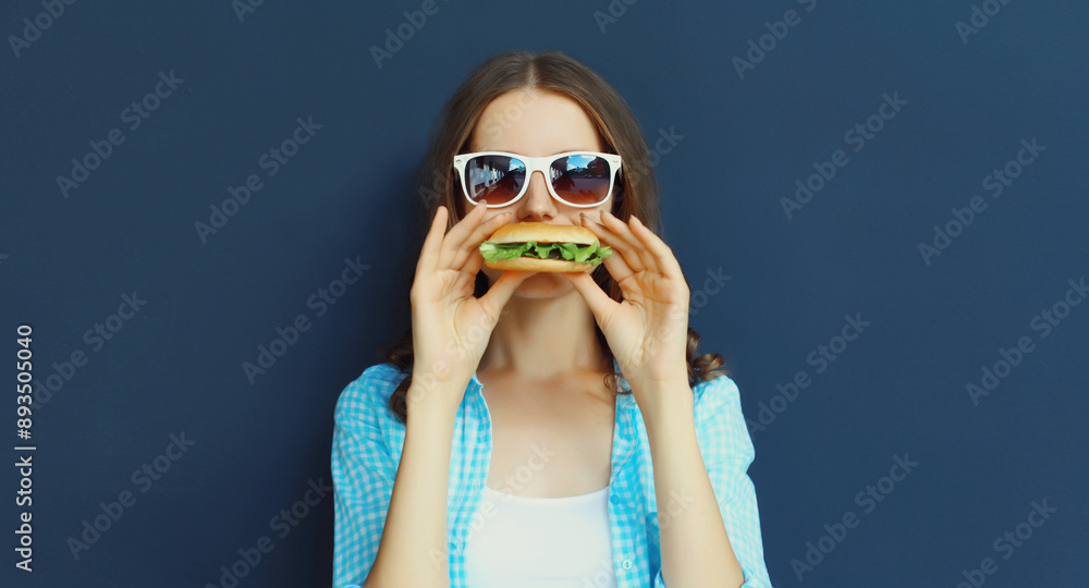 Wall mural Portrait of happy modern young woman eating burger fast food isolated on black wall background