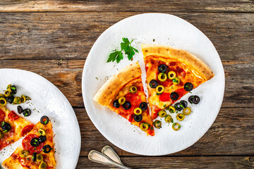 Circle pizza chorizo slices with black and green olives and mozzarella served on white plate on wooden table 
