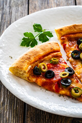 Circle pizza chorizo slices with black and green olives and mozzarella served on white plate on wooden table 