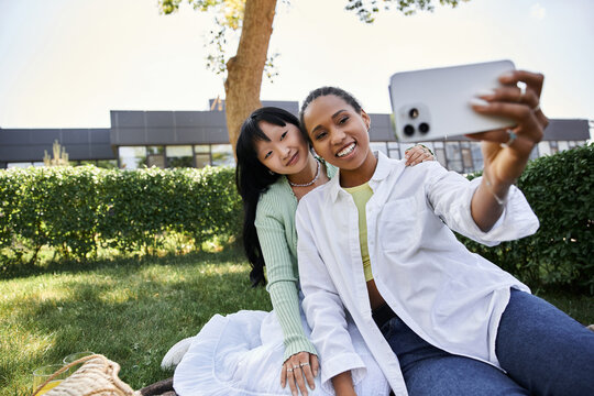 Two young women, one African American and one Asian, are sitting on a blanket in a grassy park, taking a selfie with a smartphone.