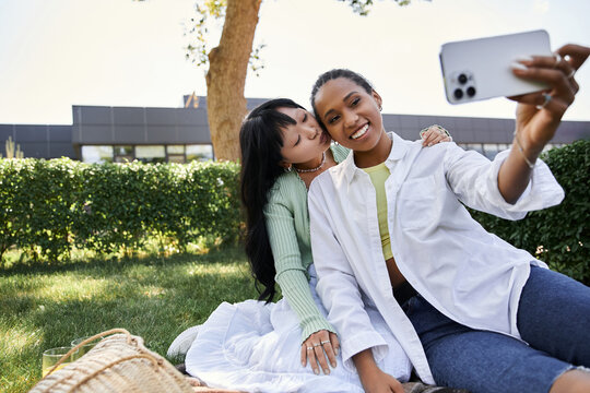 Two young women, one African American and one Asian, are on a picnic in a park, taking a selfie while sharing a tender moment.