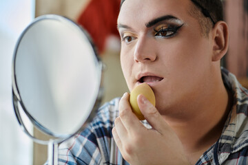 A queer person applies makeup while looking in the mirror.