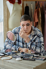 A queer person applies makeup while looking in the mirror, surrounded by an array of beauty tools.