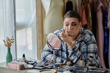 A queer person applies makeup while looking in a mirror, surrounded by makeup supplies.