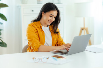 Positive successful gorgeous brunette arabian or indian ethnicity young woman, IT specialist, product manager sits at a work desk working in a laptop in office, looking device screen, smile happily
