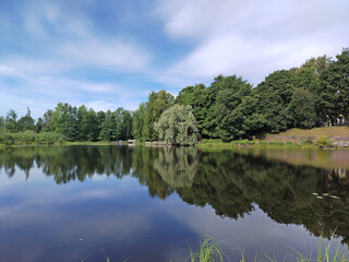 A blue lake on the shore has green trees that are reflected in the water, on a sunny day with a blue sky and white clouds.