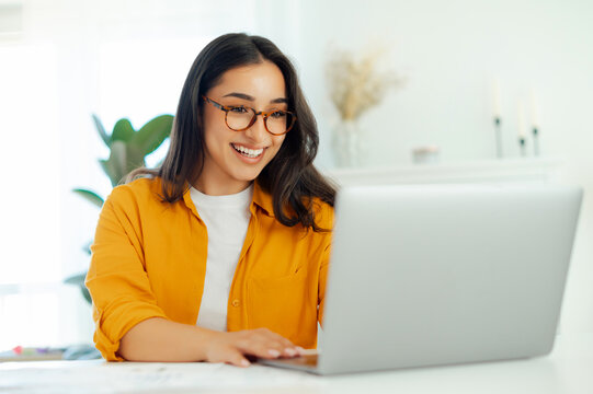 Happy successful gorgeous arabian or indian ethnicity young woman with eyeglasses, company seo, creative manager, IT specialist sits at a work desk working in a laptop, looking device screen, smile