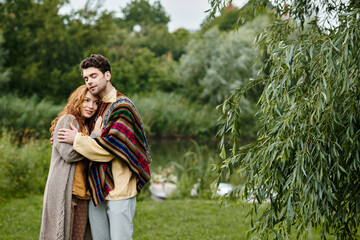 A man and a woman in boho style clothes stand closely together in a green park, exuding an air of romance and elegance.