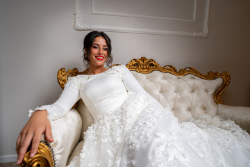 A beautiful black hair bride in a wedding dress with make-up with and jewelry sitting on a sofa 