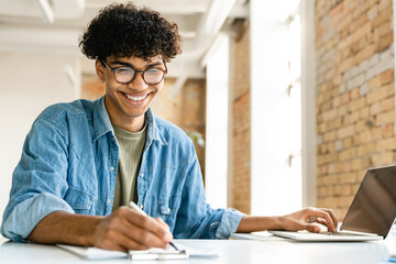 Confident smiling young mixed-race business man guy freelancer student working remote distant studding doing project homework tasks at the office desk while sitting in modern loft cafe coworking