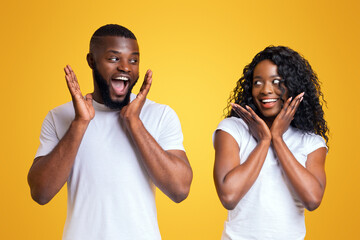 A man and a woman are standing in front of a yellow background, both looking shocked and with their hands on their cheeks.