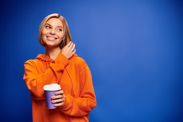 appealing joyous woman with short blonde hair in vibrant clothes holding coffee cup on blue backdrop