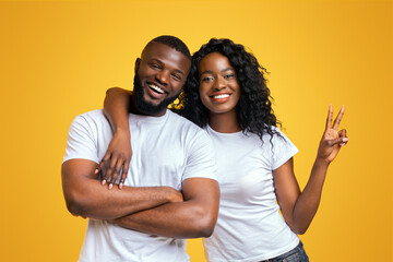 Fototapeta premium Attractive Black Girl Is Embracing Her Guy And Showing Peace Gesture On Yellow Background