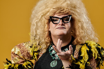 A queer person in a glamorous outfit poses against a yellow background.