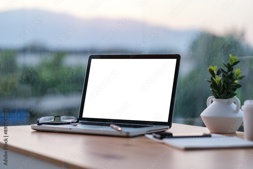 Poster laptop with a blank white screen is placed on a wooden desk in front of a large window, providing a 