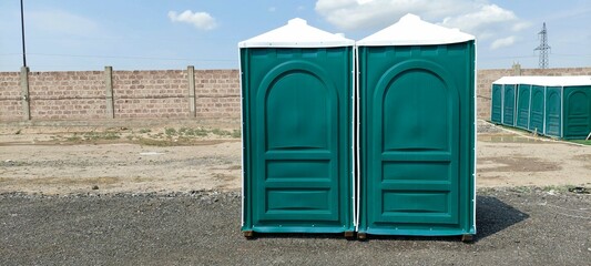 Two green portable toilets standing on a gravel surface outdoors against a stone wall with blue sky in the background, emphasizing outdoor facilities, clean design, and utility.