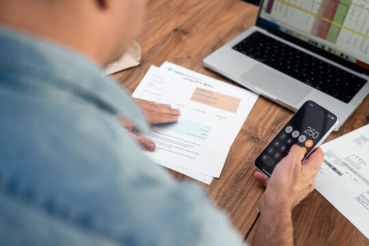 Man using a smartphone calculator to review utility bills at a wooden table. Financial planning and household budgeting concept. Ideal for expense management and personal finance topics. 