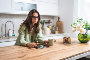 Smiling young woman wearing glasses drinking coffee and using tablet computer while sitting in kitchen. Businesswoman reading emails at morning in her home. Remote work business female