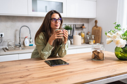 Smiling young woman wearing glasses drinking coffee and using tablet computer while sitting in kitchen. Businesswoman reading emails at morning in her home. Remote work business female