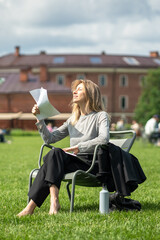 Exhausted and displeased woman squints in the bright sun, fans herself with papers, sprawls in...