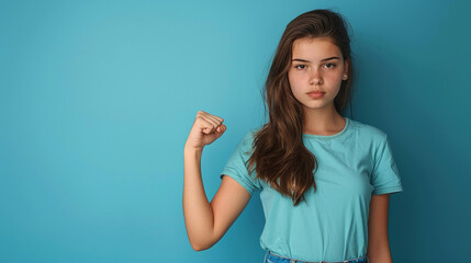 Fototapeta premium A teenage girl in a blue shirt, standing against a matching blue background, making a fist with a determined expression, symbolizing strength and determination.