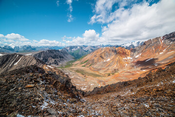 Top view from abyss edge between rocks to multicolor valley of iron colors and big sharp rocky ridge of red color. Colorful large mountains in freshly fallen snow in low clouds. Vivid alpine scenery.