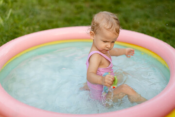 Baby in pink swimsuit is climbing out of an inflatable pool on the grass in the backyard. 