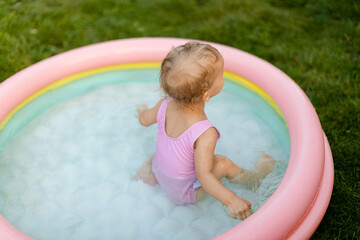Baby in a pink swimsuit is swimming in inflatable pool in the backyard. No face. Back view. 