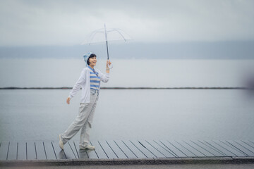 Naklejka premium Side view of young Asian woman walking and holding umbrella with lake background, In the rainy season concept.