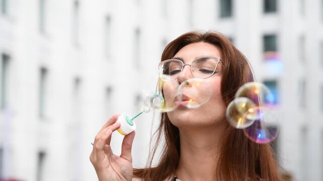 Close-up portrait of beautiful long haired brunette woman in eyeglasses and leopard print dress blowing soap bubbles on city street with white buildings in a sunny day. Soft focus. Summer leisure
