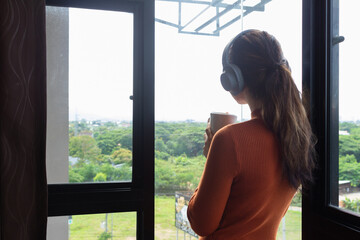 woman holds hot coffee cup in her hand and drinks it while looking at beautiful scenery outside window of vacation home. Young Asian woman drinking hot coffee by window looking at beautiful scenery