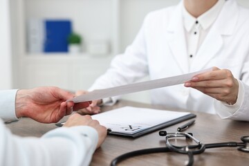 Doctor giving prescription to patient at wooden table in clinic, closeup