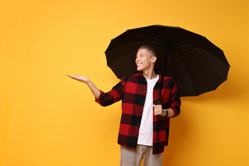 Young man with black umbrella on yellow background, space for text