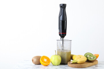 Hand blender with mixture of ingredients and fresh fruits on white marble table, space for text