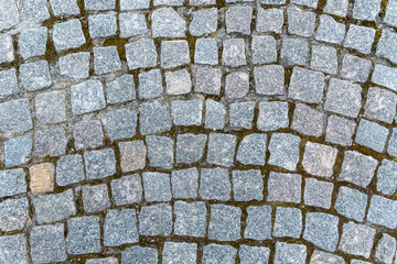 Natural stone background and texture in gray color close up. Pavement paved with square and uneven stones. Paving street with pattern. Between crushed cobblestones there are earth sand and grass.