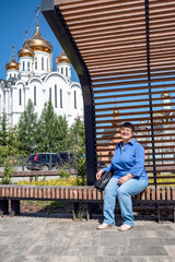 An elderly woman sits on a bench against the backdrop of a white Orthodox church with golden bathers.