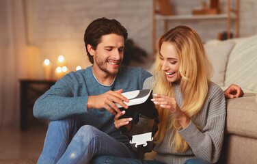 Boyfriend Giving Virtual Reality Headset To Happy Girlfriend Sitting On Floor At Home In The Evening. Selective Focus
