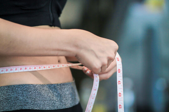 A young woman holds a tape measure to measure her waist circumference after exercising to reduce belly fat and measures her waist circumference to check the results of the weight loss course.