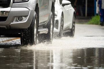 A white car is driving in the rain and splashing water on a grey car. The scene is wet and rainy, and the cars are driving on a wet road