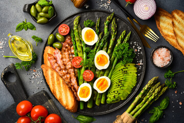 Tasty breakfast. Sunny side up eggs, asparagus, bacon and avocado. On a black stone plate.